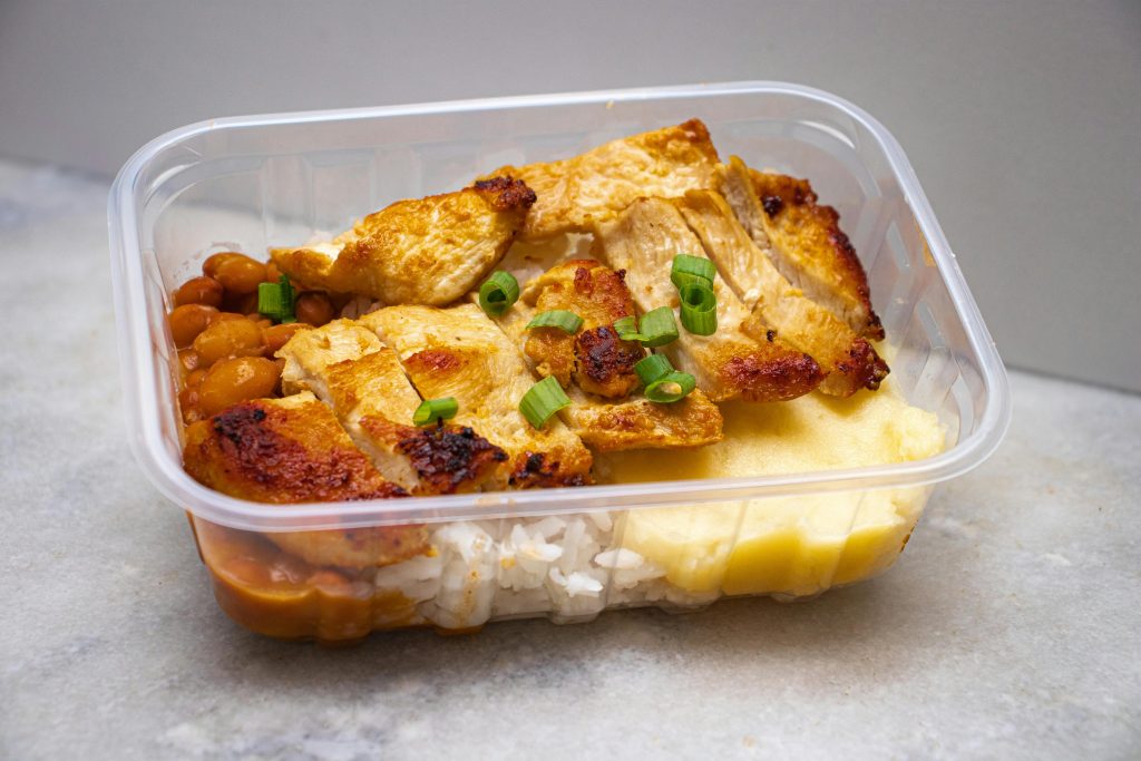 pexels-photo-30635708-30635708 Grilled chicken with rice, beans, and greens in a clear takeout container.